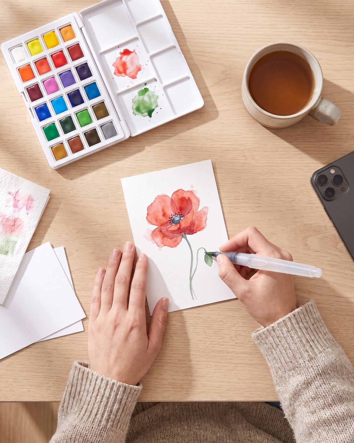 Hands finishing a watercolor poppy on a postcard with the Tintlet water brush, palette open beside it